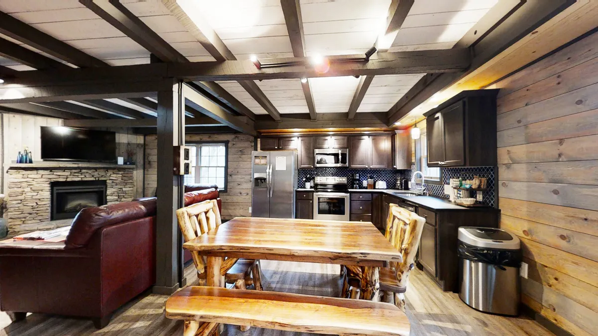 This image captures a warm open-concept space featuring a rustic wooden dining table, comfortable leather sofa, modern kitchen appliances, and a stone fireplace in the background. The arrangement emphasizes communal gathering, with each area of the living, dining, and kitchen spaces clearly visible and seamlessly interconnected.