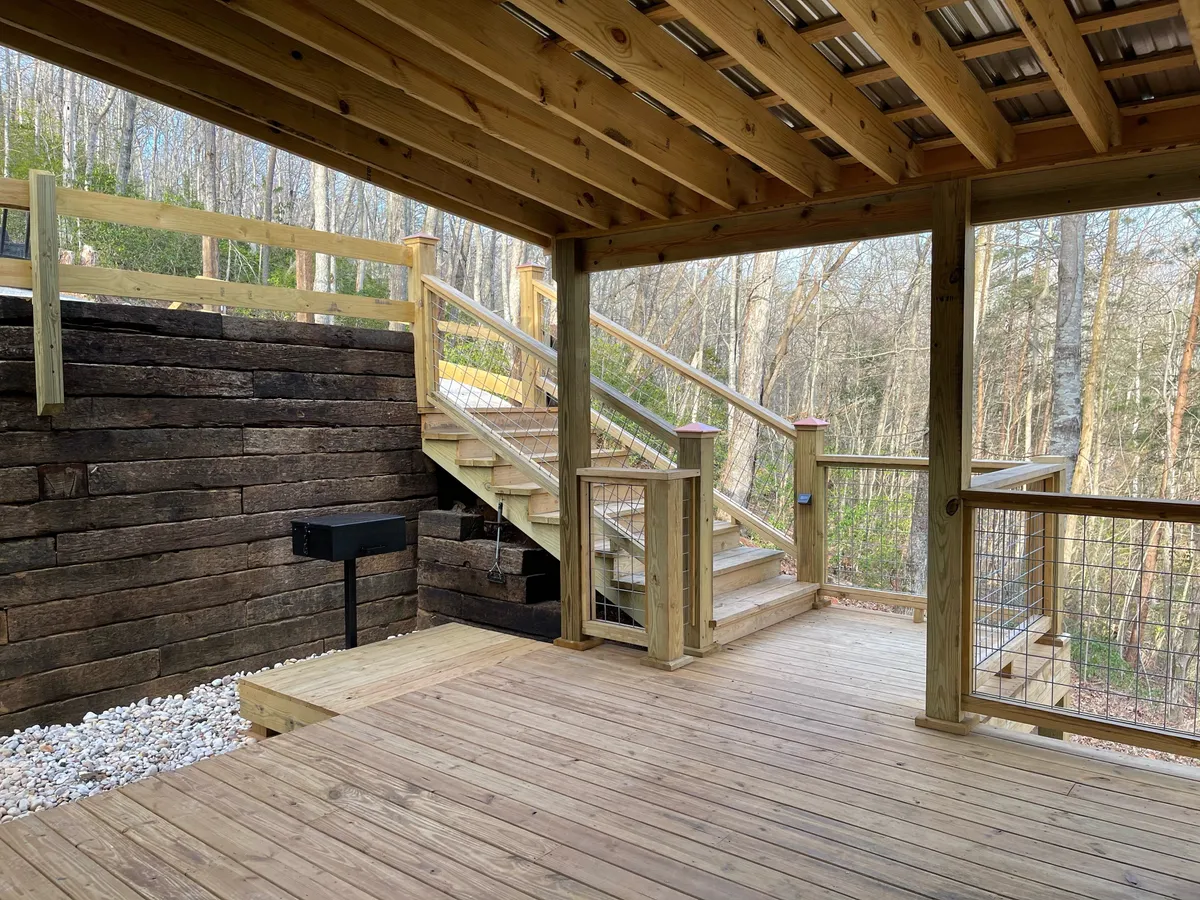 This exterior image focuses on the spacious wooden deck, highlighting a charcoal grill ready for outdoor cooking, with a stone wall and inviting space for meals or gatherings. The staircase and natural surroundings add to the sense of openness and connection with the outdoors.