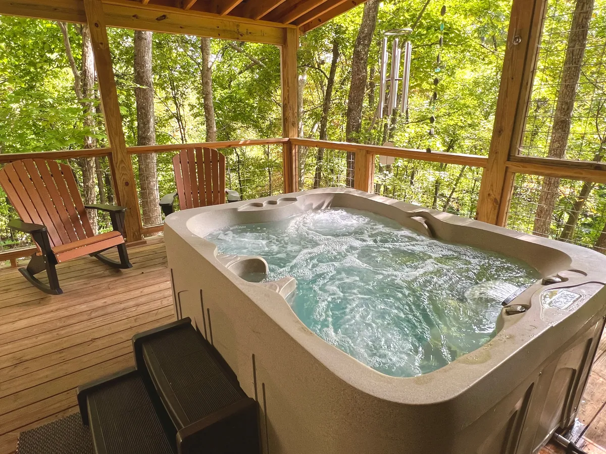 A peaceful porch scene featuring a bubbling hot tub, wooden chairs, and a view of natural surroundings. The atmosphere is inviting, with the option to relax in the hot tub under the stars or enjoy coffee on the porch while socializing with family and friends.
