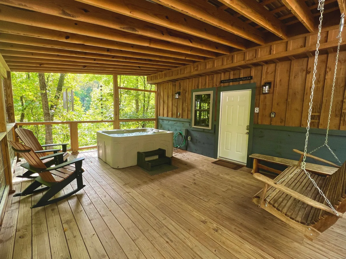 An inviting cabin deck with a bubbling hot tub, a cozy swing chair, and Adirondack chairs set on wooden flooring. The space overlooks natural scenery, suggesting opportunities for both relaxation and socializing after a day of adventure.