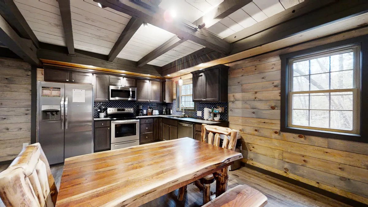 A spacious dining area is highlighted, centered around a large wooden dining table, surrounded by chairs and complemented by stainless steel kitchen appliances and a black-and-white tiled backsplash. The visible ceiling beams and overall design evoke warmth and hospitality, perfect for shared meals and conversations.