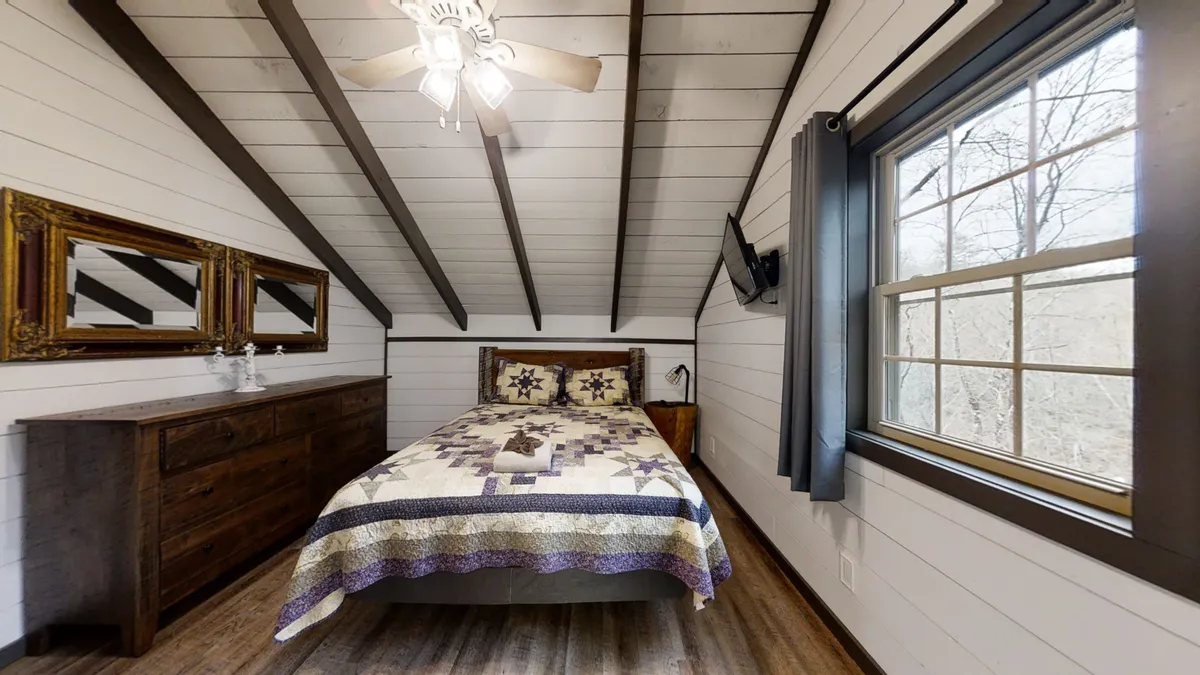 This image features a cozy bed adorned with a quilt, a wooden dresser, and a large mirror. Natural light streams into the room, creating an inviting and serene atmosphere ideal for relaxation. The layout showcases key bedroom amenities and a tranquil retreat.