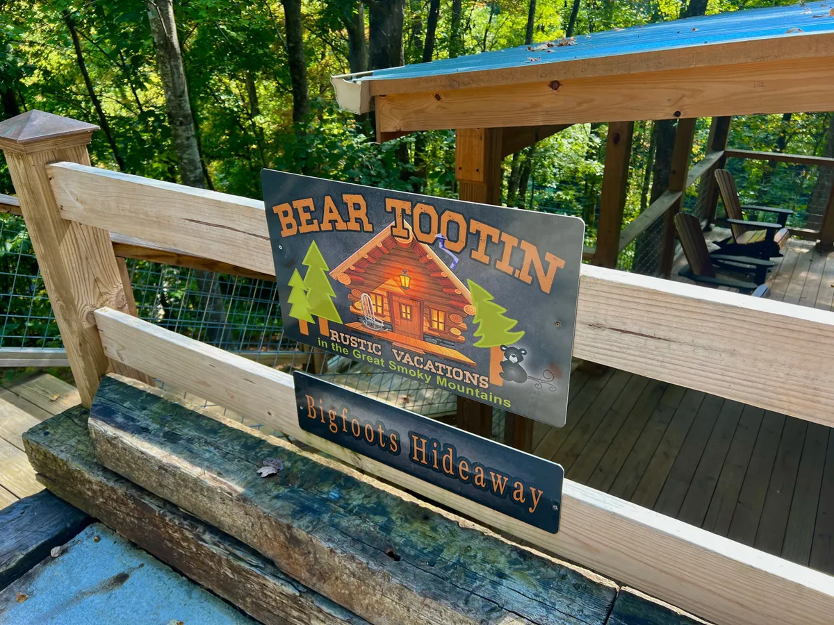 A welcoming exterior shot of the cabin surrounded by lush trees, featuring a rustic wooden sign and a spacious deck area. The image captures the cabin's playful and adventurous character, set against a backdrop of natural beauty, ideal for family gatherings and outdoor fun.