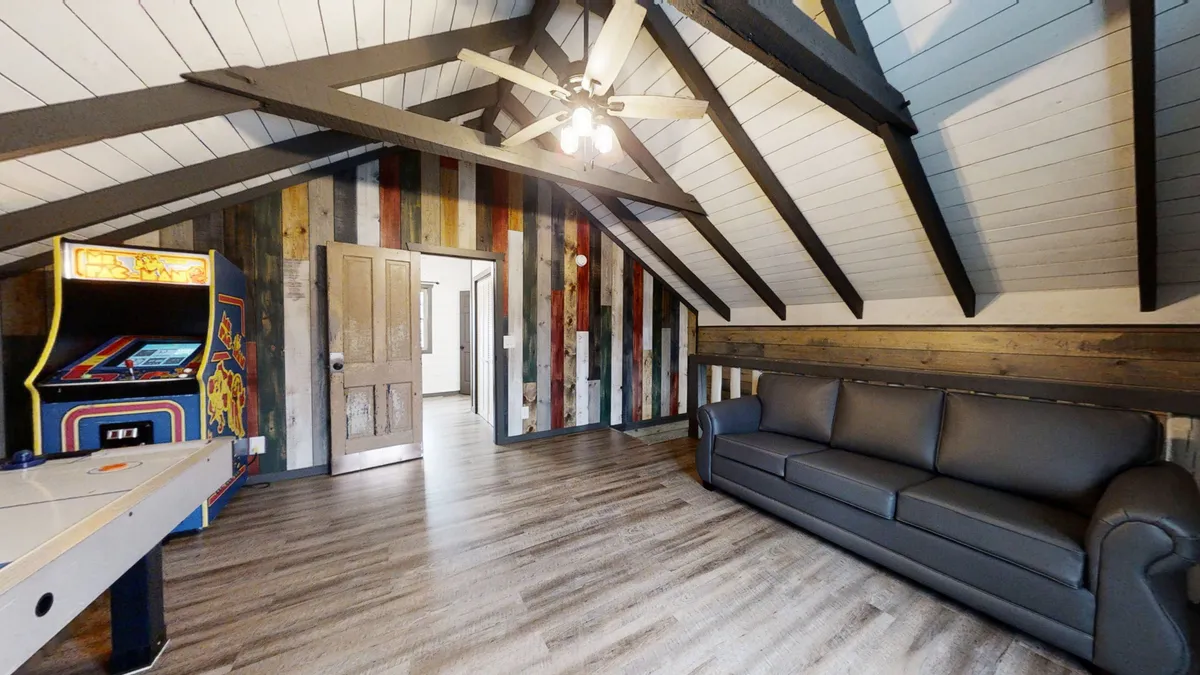This image captures the lively game loft with a clear focus on the arcade game machine, air hockey table, sofa, wooden beams, and a vibrant accent wall. The space is set up for both gaming and relaxation, with ample seating for socializing and natural light filtering in.