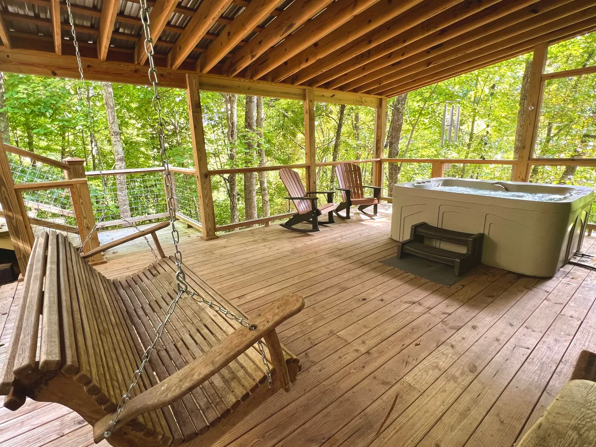 A peaceful wooden deck surrounded by lush trees features a swinging bench and a private hot tub. The swing faces outward toward nature, and the hot tub is perfectly situated for enjoying the view. The deck exudes a cozy, intimate ambiance, ideal for unwinding and connecting with nature.