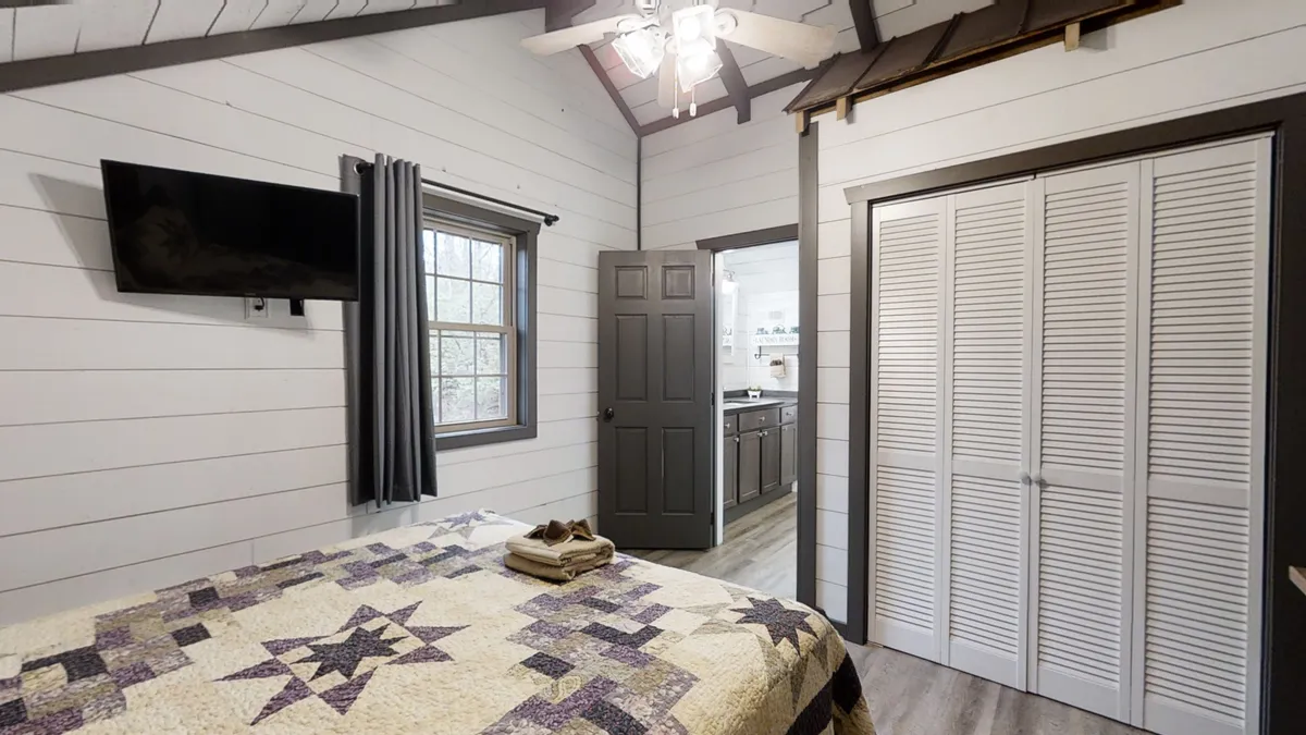 The photo presents a quilted bedspread, a window with curtains, a TV mounted on the wall, and closed closet doors. The setting feels both comfortable and homey, emphasizing relaxation and modern convenience within the master suite.