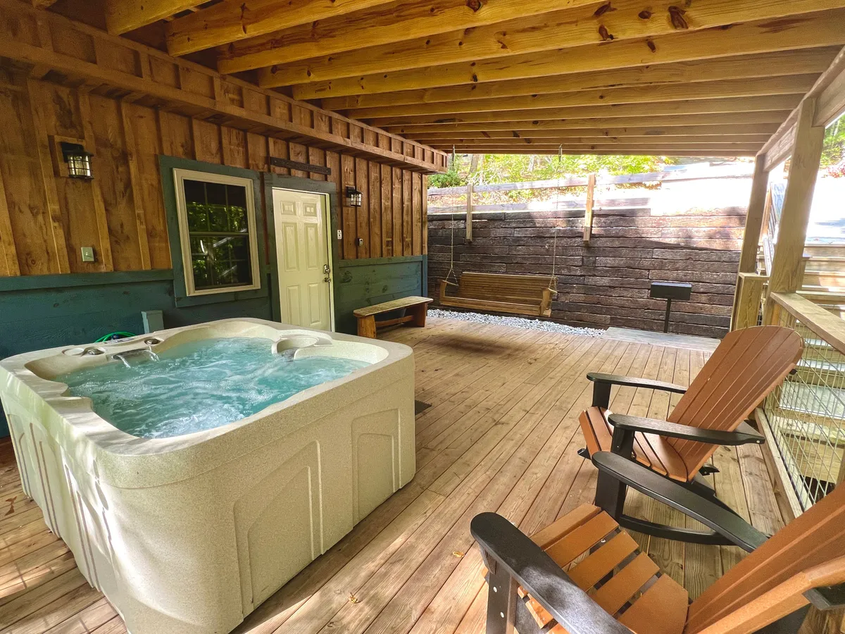 An inviting deck set among the trees features a bubbling outdoor hot tub, a classic wooden swing, and Adirondack chairs perfect for relaxing. The area is bathed in soft natural light, suggesting opportunities for stargazing or sunset views from the comfort of the deck.