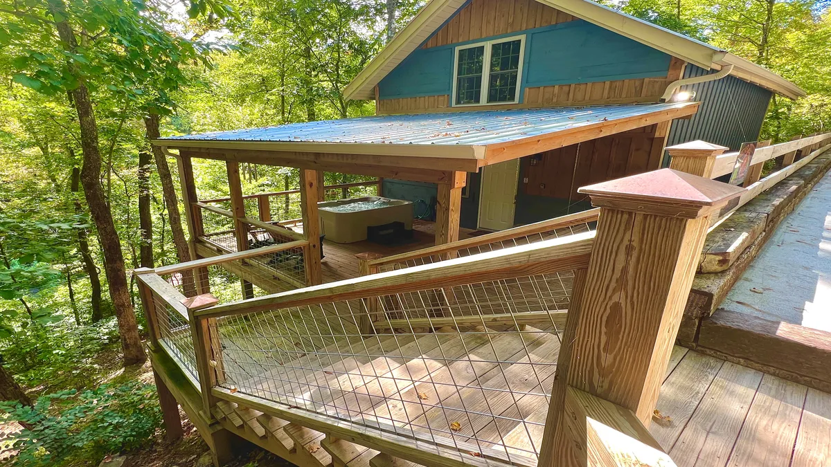 The image showcases the rustic blue exterior of Bigfoot's Hideaway cabin, with a spacious wooden deck extending into the surrounding lush Smoky Mountain forest. The scene is bathed in peaceful sunset light, inviting guests to relax outdoors, enjoy the tranquil scenery, and connect with nature.