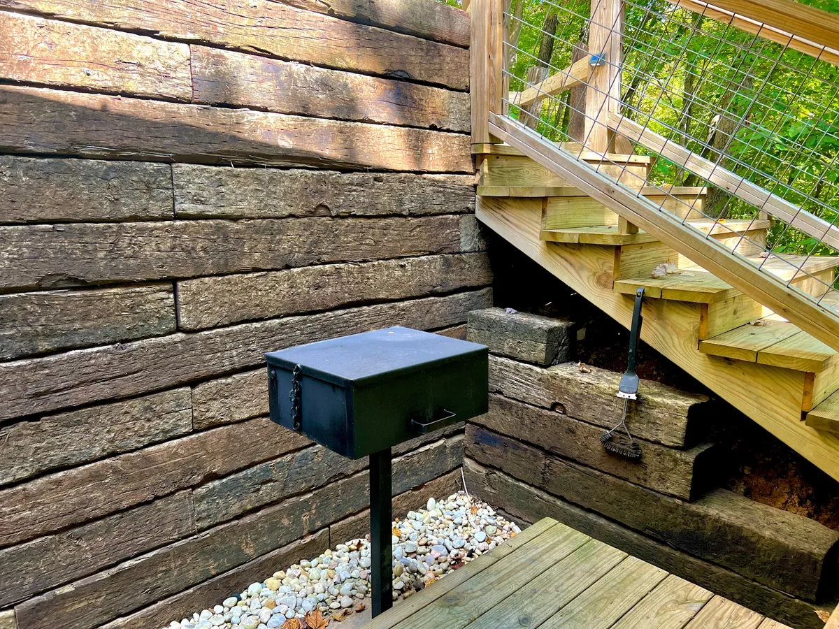 This exterior image features a welcoming cabin entrance with a black mailbox, wooden stairs, and a rustic stone wall. The setting hints at outdoor gathering possibilities and the tranquil, natural surroundings guests can expect when arriving for a group getaway.
