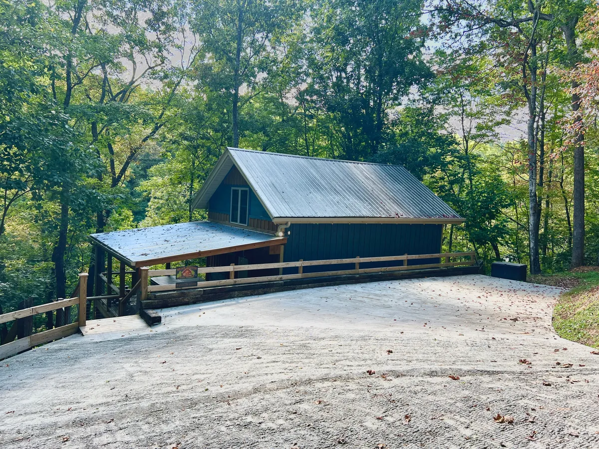 A serene view of the cabin nestled among trees, with a gravel driveway leading up to it. The porch invites guests to relax, while the natural surroundings evoke the sense of being steps away from adventure in the Smokies.